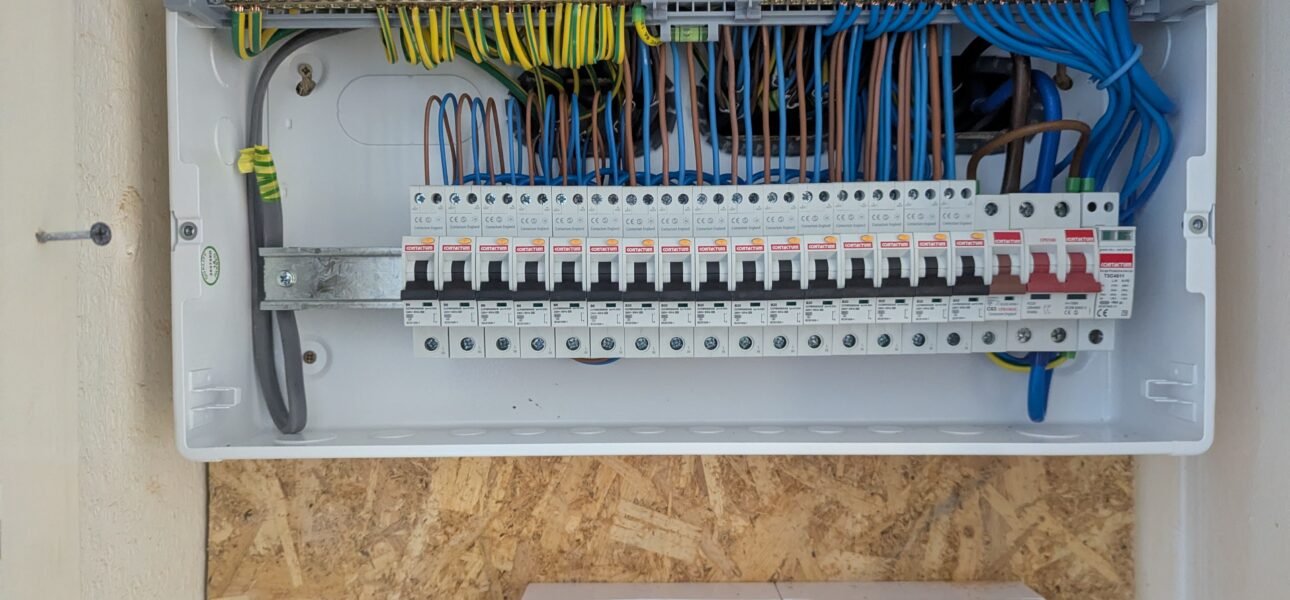 A domestic consumer unit (fuseboard) with multiple MCBs and RCDs installed, showing trunking with colour-coded cables — brown live, blue neutral, and green/yellow earth — alongside two white surface-mounted double sockets on a timber-backed wall.