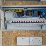 A domestic consumer unit (fuseboard) with multiple MCBs and RCDs installed, showing trunking with colour-coded cables — brown live, blue neutral, and green/yellow earth — alongside two white surface-mounted double sockets on a timber-backed wall.